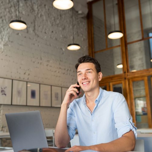 modern young handsome man sitting in co-working office, talking on phone, casual style, confident, working on kaptop online, freelancer, busy